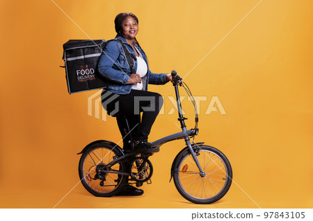 Portrait of fast food delivery worker riding bike during lunch time while carrying thermal takeout backpack, deliverying meal order to customer. Food service and transportation 97843105
