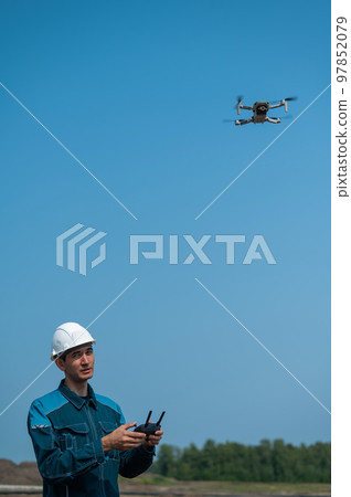 A man in a helmet and overalls controls a drone at a construction site. The builder carries out technical oversight. 97852079