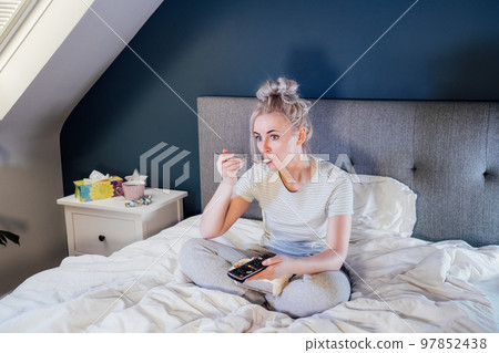 Frustrated woman in home clothes eating ice cream, using the remote control to change the channel while glazing TV in bed during day. Overeating, Jamming of negative emotions. Selective focus. 97852438