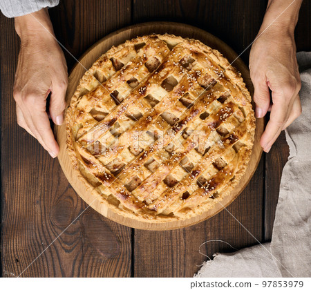 Two female hands hold a round baked pie with apple filling on a wooden board, brown table. View from above 97853979