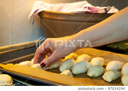 A woman's hand puts small raw pies in a pan. Meat pies are traditional Latvian holiday dishes. 97854294