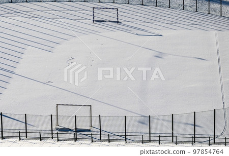 Winter snow-covered football field and frosty sunny weather. Long shadows fall from the fencing of a football pitch into the snow. Winter snow-covered football field and frosty sunny weather. Long shadows fall from the fencing of a football pitch into the snow. 97854764