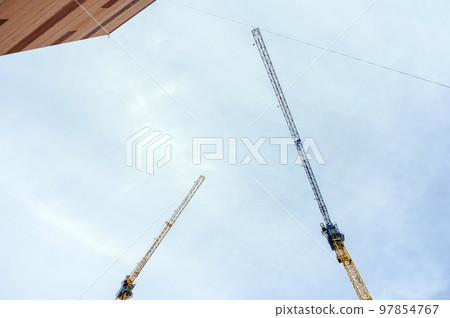 Two long arrows of large tower construction cranes in blue and yellow against a bright sunny sky. Near the wall of a high-rise brick house. Two long arrows of large tower construction cranes in blue and yellow against a bright sunny sky. Near the wall of a high-rise brick house. 97854767