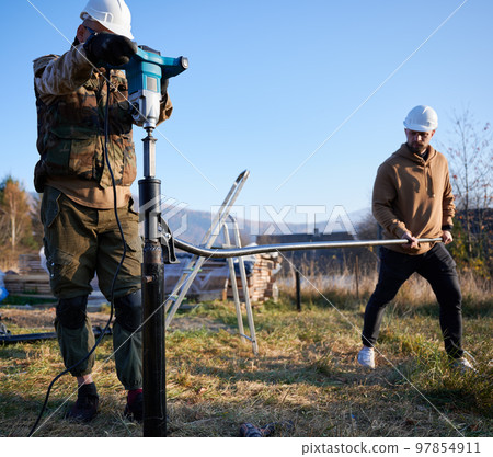 Male workers building pile foundation for wooden frame house. Men builders in white safety helmets drilling piles into the ground on blue sky background. 97854911