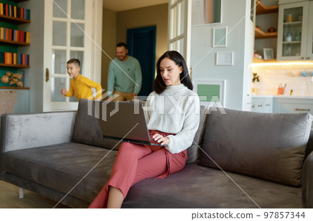 Young mother trying to work on laptop online while father and son playing 97857344