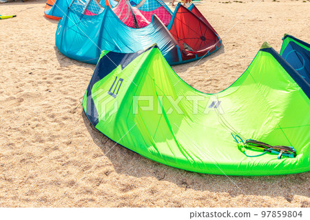 Close-up many bright kitesurf wings and kite equipment sand beach shore watersport spot on bright sunny day against sea coast blue sky and kiter riding surf school camp. Active travel sport concept Close-up many bright kitesurf wings and kite equipment sand beach shore watersport spot on bright sunny day against sea coast blue sky and kiter riding surf school camp. Active travel sport concept 97859804