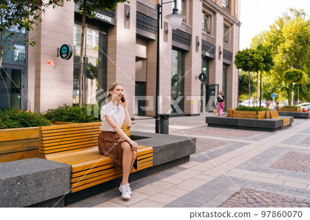 Wide shot of elegant young woman with blond hair having mobile phone conversation sitting on bench on city street in summer day. Focused lady making call on mobile phone, speaking about work issues. 97860070