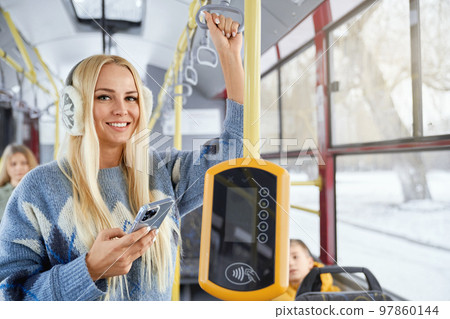 Portrait of fair long haired pretty female smiling nicely and looking directly in camera holding smarphone in one hand performing contactless payment and other one grasping holder with earmuffs. Portrait of fair long haired pretty female smiling nicely and looking directly in camera holding smarphone in one hand performing contactless payment and other one grasping holder with earmuffs. 97860144