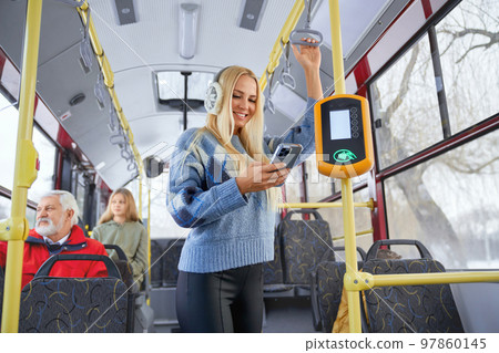 Portrait of fair long haired female wearing blue sweater and black trousers with earmuffs standing near bus payment terminal and performing contactless operation passengers watching city from window. 97860145