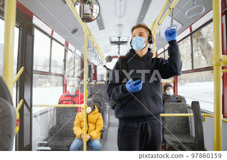 Front view of passengers going home by public transport, standing, sitting, wearing medical masks and gloves. Young man holding smartphone, paying. Concept of urban life. 97860159