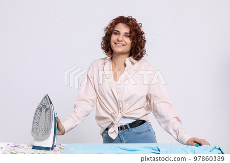 The girl smiles gratefully while standing by the ironing board and iron. The girl smiles gratefully while standing by the ironing board and iron. 97860389