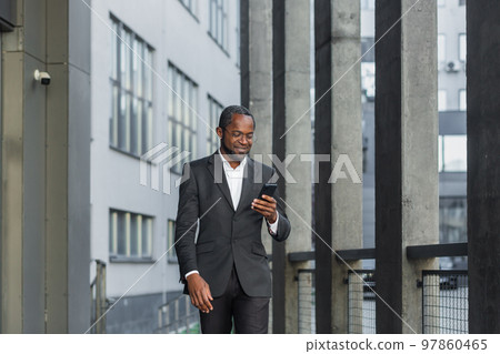 Happy successful African American businessman boss walking outside office building, manager holding smartphone reading message and smiling, using communication app. Happy successful African American businessman boss walking outside office building, manager holding smartphone reading message and smiling, using communication app. 97860465