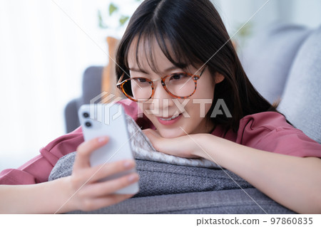 Young woman with glasses lying on the sofa and using a smartphone Young woman with glasses lying on the sofa and using a smartphone 97860835