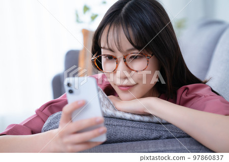 戴眼鏡的年輕女子躺在沙發上使用智能手機 戴眼鏡的年輕女子躺在沙發上使用智能手機 97860837