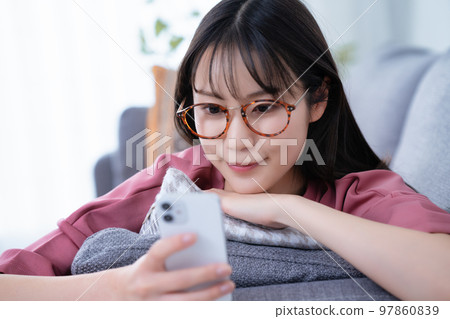 戴眼鏡的年輕女子躺在沙發上使用智能手機 97860839