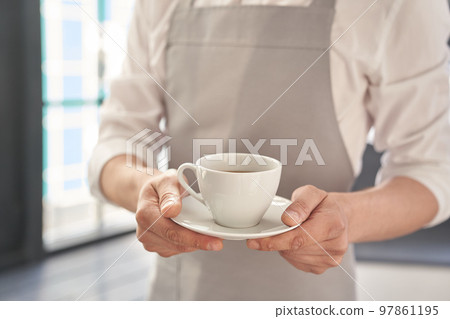 Hands of the waiter in a gray apron holds out a cup of coffee. Barista gives a cup of hot coffee in a cafe, against the background of a large window. High quality photo 97861195