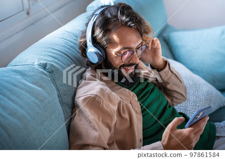 Smiling young man wearing headphones listen music playing in smartphone sitting on sofa at home Smiling young man wearing headphones listen music playing in smartphone sitting on sofa at home 97861584