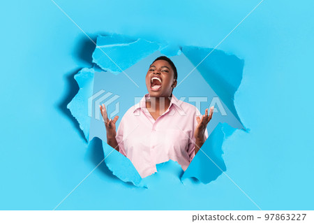 Angry oversize black woman shouting freaking out posing in hole of torn paper on blue studio background 97863227