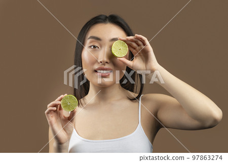Fruit acids for skin care. Beautiful asian lady holding lime halves in front of her face on brown studio background Fruit acids for skin care. Beautiful asian lady holding lime halves in front of her face on brown studio background 97863274