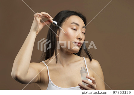 Beautiful asian woman applying face serum with dropper, enjoying new beauty product, brown background Beautiful asian woman applying face serum with dropper, enjoying new beauty product, brown background 97863339