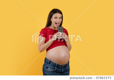 Portrait Of Cheerful Young Pregnant Lady Biting Chocolate Bar Over Yellow Background 97863373