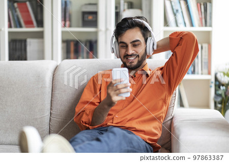 Happy arab guy relaxing on couch with phone and headphones 97863387