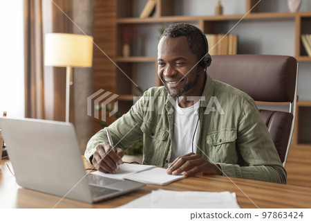 Smiling mature black male manager in headphones watch video on computer, makes notes at workplace 97863424