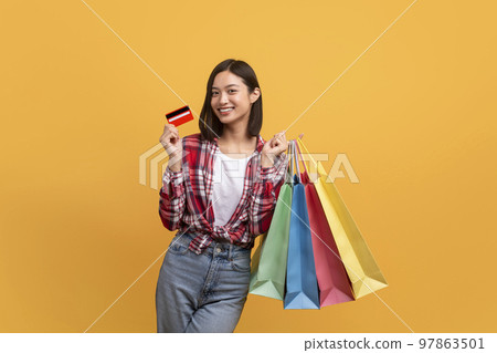 Cashback concept. Happy asian lady posing with credit card and colorful shopping bags over yellow studio background 97863501