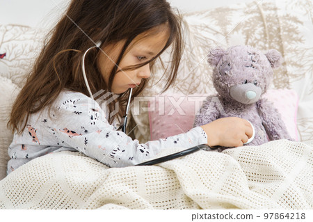 Serious little girl in sleepwear sitting under blanket and listening to lung of teddy bear with stethoscope. Bed rest. Serious little girl in sleepwear sitting under blanket and listening to lung of teddy bear with stethoscope. Bed rest. 97864218