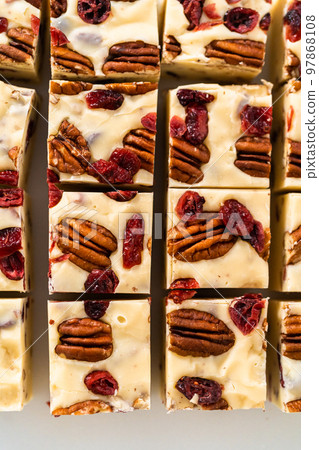 Cutting white chocolate cranberry pecan fudge into small pieces on a white cutting board. Cutting white chocolate cranberry pecan fudge into small pieces on a white cutting board. 97868108
