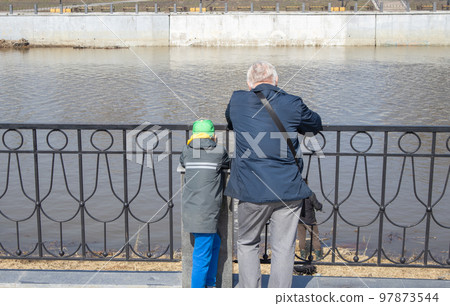 A gray-haired grandfather and his grandson are standing on the river embankment and leaning on metal handrails, looking at a fisherman on a sunny spring day A gray-haired grandfather and his grandson are standing on the river embankment and leaning on metal handrails, looking at a fisherman on a sunny spring day 97873544