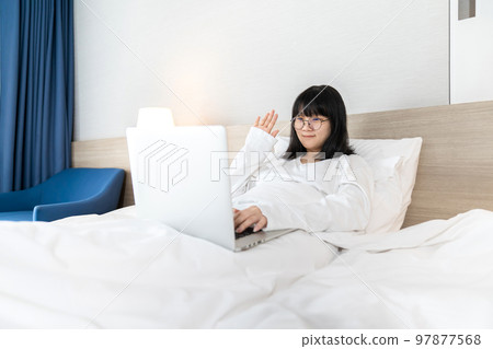 Glasses Asian young pretty teenager rise hand up hi and hello to laptop notebook for meeting and video conference on the bed in Work From Home period. Glasses Asian young pretty teenager rise hand up hi and hello to laptop notebook for meeting and video conference on the bed in Work From Home period. 97877568