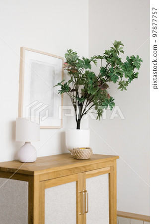 An artificial flower in a pot on a wooden chest of drawers in the living room, decorative items and a frame on the wall in a Scandinavian minimalist style 97877757