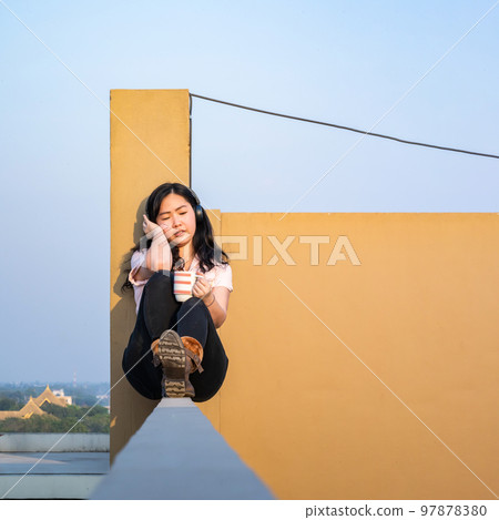 Glasses Asian woman is listening music via headset, drinking coffee on rooftop barricade cement (not dangerous because the opposite site is rooftop floor as well). Glasses Asian woman is listening music via headset, drinking coffee on rooftop barricade cement (not dangerous because the opposite site is rooftop floor as well). 97878380