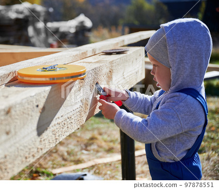 Boy toddler playing as builder on construction site. Child carpenter in blue overalls learning to build wooden frame house outdoor on sunny day. Carpentry and workshop concept. 97878551