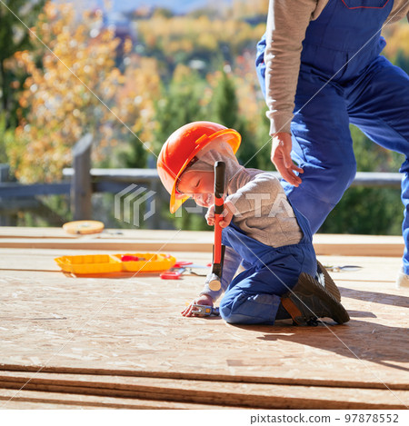 Father with toddler son building wooden frame house. Male builders hammering nail into plank on construction site, wearing helmet and blue overalls on sunny day. Carpentry and family concept. 97878552