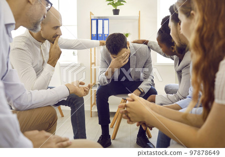 Sad man at support group therapy session sitting with head down and participants touch shoulders. 97878649
