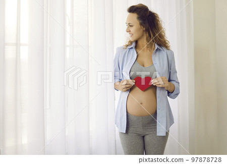 Smiling beautiful future young mother holding red heart near big pregnant belly. Smiling beautiful future young mother holding red heart near big pregnant belly. 97878728