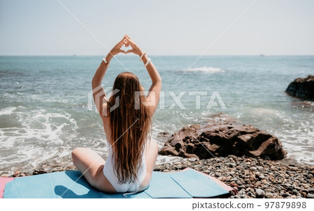 Woman makes heart with hands on beach. Young woman with long hair in white swimsuit and boho style braclets practicing by sea ocean. Concept of longing daydreaming love peace contemplation vacations Woman makes heart with hands on beach. Young woman with long hair in white swimsuit and boho style braclets practicing by sea ocean. Concept of longing daydreaming love peace contemplation vacations 97879898