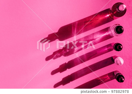 Different cosmetic glass bottles standing on viva magenta pink background. Abstract pattern by shadows and reflections in the sunlight. Red, green and yellow shades. Flat lay style with copy space 97882978