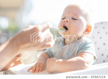 Mother spoon feeding her baby boy child in baby chair with fruit puree on a porch on summer vacations. Baby solid food introduction concept Mother spoon feeding her baby boy child in baby chair with fruit puree on a porch on summer vacations. Baby solid food introduction concept 97887219