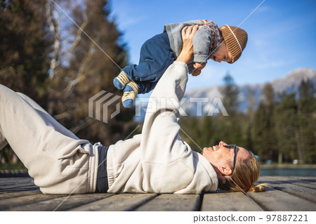 Happy family. Young mother playing with her baby boy infant oudoors on sunny autumn day. Portrait of mom and little son on wooden platform by lake. Positive human emotions, feelings, joy Happy family. Young mother playing with her baby boy infant oudoors on sunny autumn day. Portrait of mom and little son on wooden platform by lake. Positive human emotions, feelings, joy 97887221