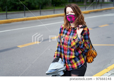 Young woman wearing a plaid handmade face mask and colorful shirt with mesh bag full of oranges and a backpack standing on the street. Young woman wearing a plaid handmade face mask and colorful shirt with mesh bag full of oranges and a backpack standing on the street. 97888148