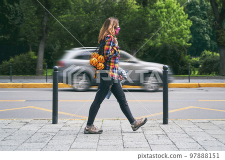 Young woman wearing a plaid handmade face mask and colorful shirt with mesh bag full of oranges walking on the street on passing car background. Zero waste concept. 97888151