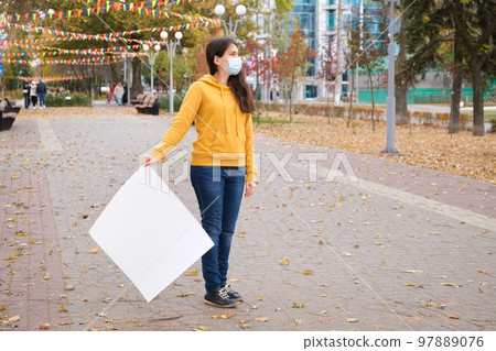 A woman in a medical mask holds an empty white sheet of paper for the inscription and looks away. 97889076