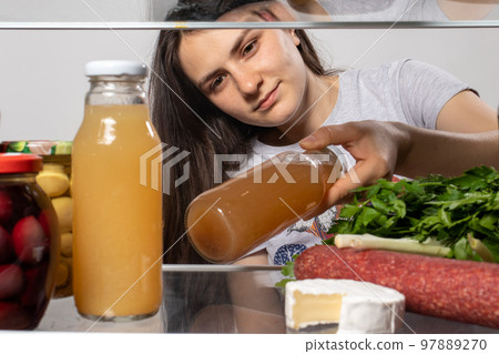 The girl looks in the fridge and takes out fruit juice in a glass bottle. Products in the refrigerator. 97889270