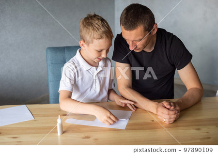 A cute boy is sitting with his dad at the table and collecting origami. Glue the parts 97890109