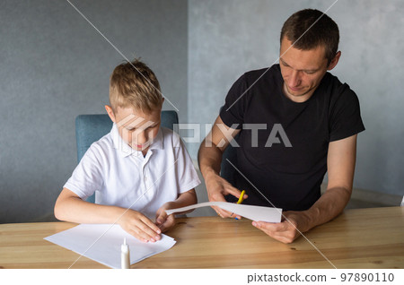 A cute boy is sitting with his dad at the table and collecting origami. Cutting a white sheet of paper A cute boy is sitting with his dad at the table and collecting origami. Cutting a white sheet of paper 97890110