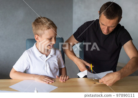 A cute boy is sitting with his dad at the table and collecting origami. Cutting a white sheet of paper 97890111
