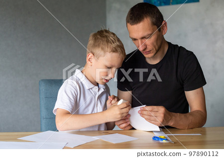 A cute boy is sitting with his dad at the table and collecting origami. Holds a white sheet of paper A cute boy is sitting with his dad at the table and collecting origami. Holds a white sheet of paper 97890112